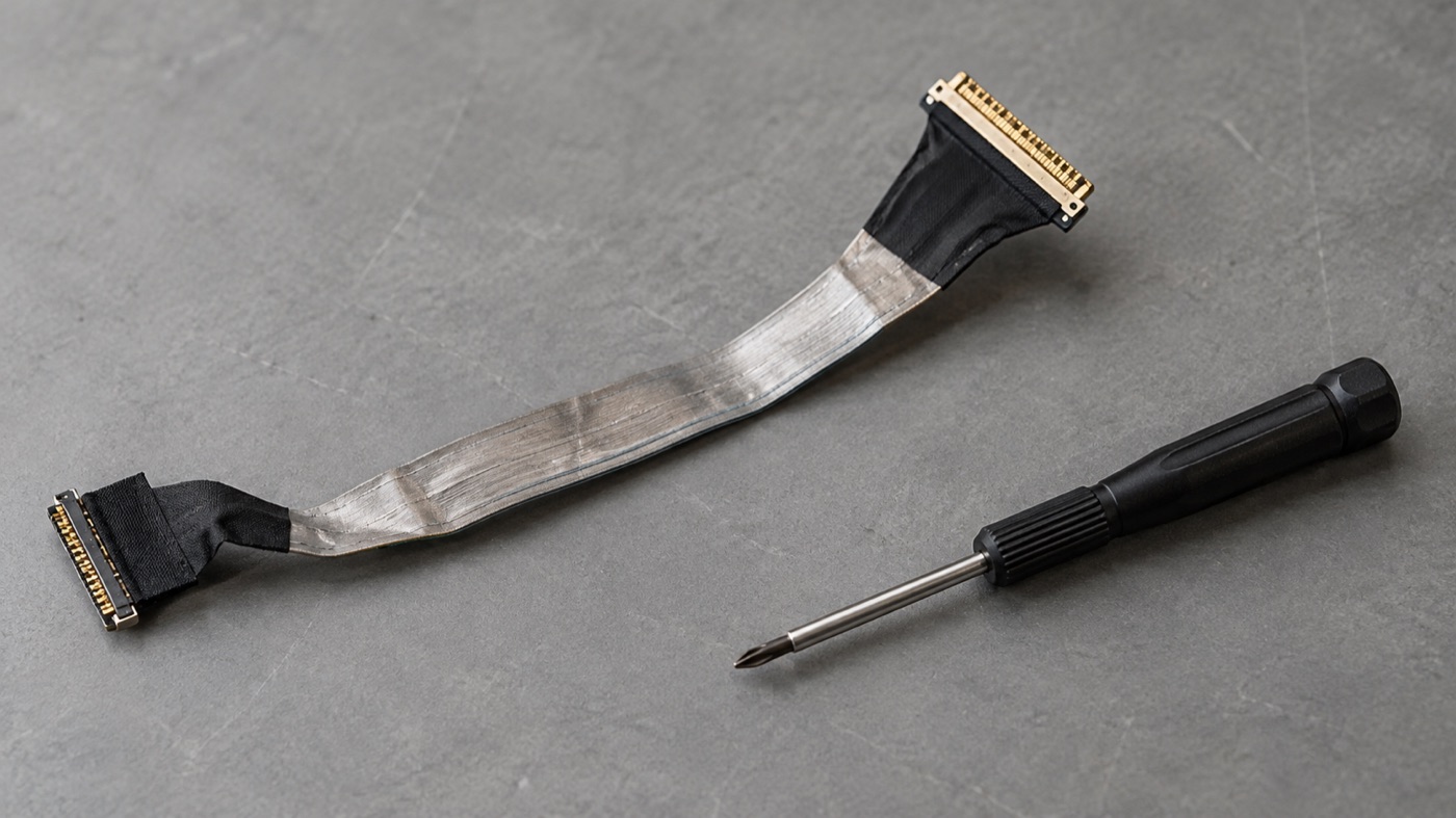 Laptop display ribbon cable and a small screwdriver on a grey workbench